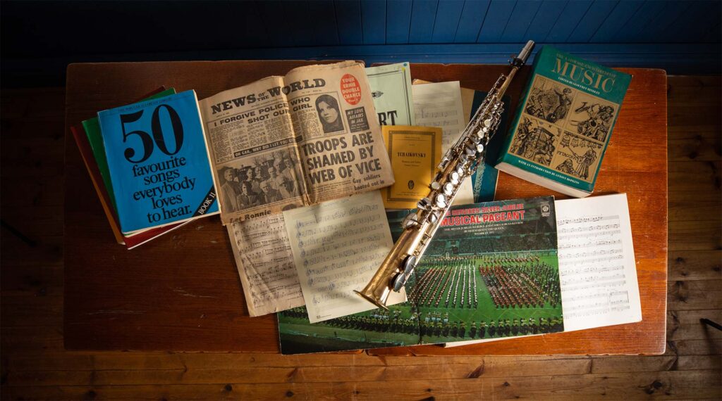 Looking directly down on a wooden table with music books, an old copy of news of the world and a wind instrument.